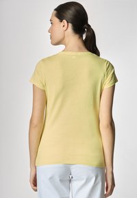Una donna con i capelli lunghi raccolti in una coda di cavallo indossa una maglietta gialla chiara, vista da dietro, su uno sfondo grigio chiaro.