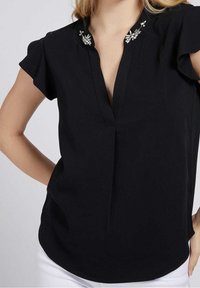 Femme portant un chemisier noir à manches courtes avec un décolleté en V profond et des ornements floraux en perles argentées sur le col.