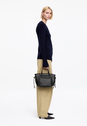 Mujer con cabello rubio corto de perfil, vestida con cárdigan azul marino, pantalones anchos beige y zapatos negros puntiagudos, sosteniendo un bolso de cuero negro por las asas.