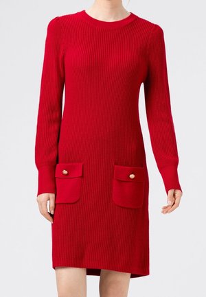 Punainen pitkähihainen neulemekko, jossa on kaksi koristeltua etutaskua kultaisilla napeilla, päällä seisovalla henkilöllä yksinkertaisen vaalean taustan edessä.