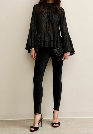 Femme portant un chemisier noir transparent à pois avec des manches cloche, un pantalon noir ajusté, des sandales à talons hauts, et tenant une pochette noire à sequins.