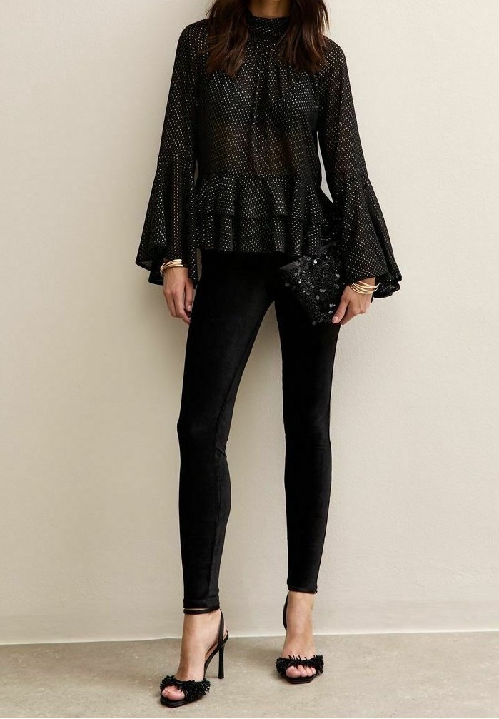 Femme portant un chemisier noir transparent à pois avec des manches cloche, un pantalon noir ajusté, des sandales à talons hauts, et tenant une pochette noire à sequins.