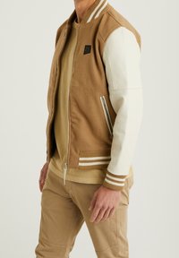 Brązowa i kremowa kurtka bomberka z wełnianym korpusem, skórzanymi rękawami i prążkowanymi mankietami. Posiada zapinaną na zamek kieszeń oraz detal z logo.
