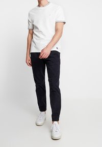 Mies, który ma na sobie biały t-shirt z krótkim rękawem, czarne dresowe spodnie oraz białe trampki, stoi na tle białego tła.