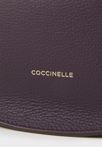 Dark purple leather with a textured surface. Features gold embossed "COCCINELLE" logo. Edge stitching visible along the perimeter.