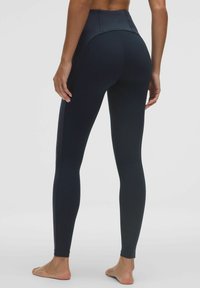 Egy fekete, magas derekú legginget viselő személy mezítláb áll, háttal a kamerának, egy sima fehér háttér előtt.