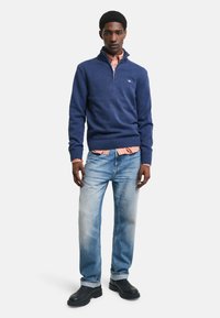 Maglione lavorato a maglia blu navy con colletto zip, indossato sopra una camicia a quadri chiari, abbinato a jeans blu sbiaditi e stivali neri.