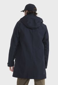 Marineblauer Regenmantel mit Kapuze, der über ein elegantes Design, strukturierten Stoff und verstellbare Manschetten verfügt. Die Rückansicht zeigt einen belüfteten unteren Teil.