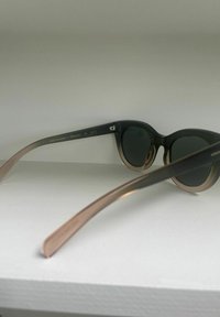 Runda solglasögon med mörka linser, en svart framram och ljusbruna genomskinliga skaft. Etikett på den inre skaftet innehåller märke och specifikationer.