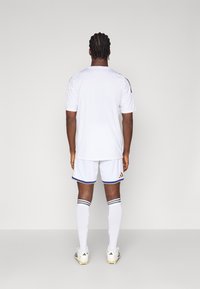 White athletic jersey with black accents, paired with white shorts featuring blue trim and gold branding, complemented by white knee-high socks.