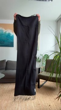 Couverture noire en matériau doux, avec des bords à franges et une texture lisse. Tenue dans une pièce avec des plantes et du mobilier moderne.