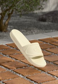 Beige Calvin Klein papucs, amelyen a lábbelin a "CK" logó és felirat van dombornyomva, fából készült blokk és kavicsos kültéri burkolat fölött elhelyezve.