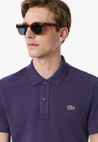 Lila polotröja med en texturerad väv, med en treknapplåstning och en liten grön och vit krokodil-logotyp på bröstet. Solglasögon med bruna bågar.