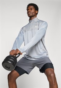 Svijetlosiva dugosavna sportska majica s zip kragnom; tamnosive kratke hlače prekrivene crnim kompresijskim hlačama; crni kettlebell u rukama.