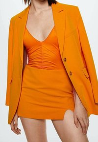 Blazer orange avec robe moulante assortie, dotée d'un décolleté en V profond et d'une fente latérale. Texture lisse, design structuré et fermeture à deux boutons.