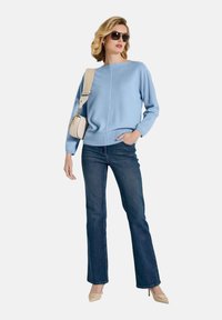 Hellblauer Pullover mit langen Ärmeln, kombiniert mit dunklen Schlagjeans und beigen High Heels, dazu eine kleine cremefarbene Schultertasche.