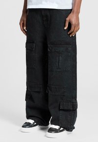 Persona che indossa ampi pantaloni cargo neri con multiple tasche a pattina e sneakers bianco e nere, in piedi contro uno sfondo semplice.