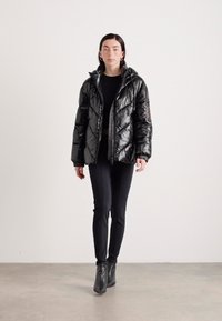 Veste noire, rembourrée, matelassée avec un capuchon et fermeture éclair. Présente des coutures diagonales et des poignets élastiques, portée avec un pantalon slim noir et des bottes.