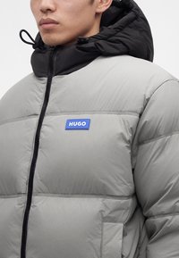 Homme portant une doudoune gris clair avec une capuche noire et un patch bleu portant la mention "HUGO" sur la poitrine gauche, fermé par une fermeture éclair.