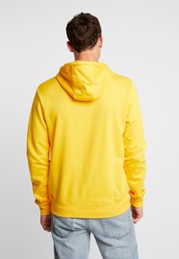 Hombre de espaldas vistiendo una sudadera amarilla brillante y pantalones vaqueros azul claro contra un fondo gris claro liso.