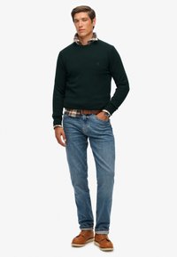 Suéter verde oscuro, camisa de cuadros debajo, jeans azules, zapatos marrones, en pose de pie. El suéter presenta un escote redondo y puños acanalados.