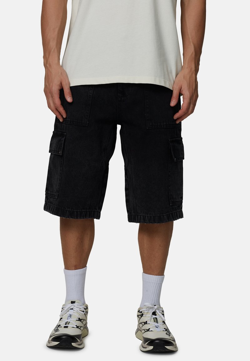 Man die zwarte cargoshorts, witte sokken, witte en zwarte sneakers en een wit T-shirt draagt, staand tegen een effen achtergrond.