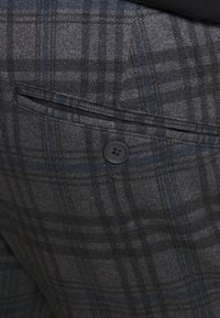 Tejido a cuadros en gris y azul con un bolsillo de ojal abotonado, mostrando una textura y costura detalladas.