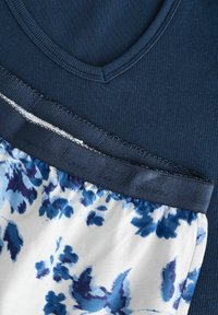 Tricou din materiale cu brâu ondulat de culoare bleumarin, suprapus peste un material alb cu imprimeu floral albastru. Prezintă un tiv ondulat și un brâu albastru contrastant.