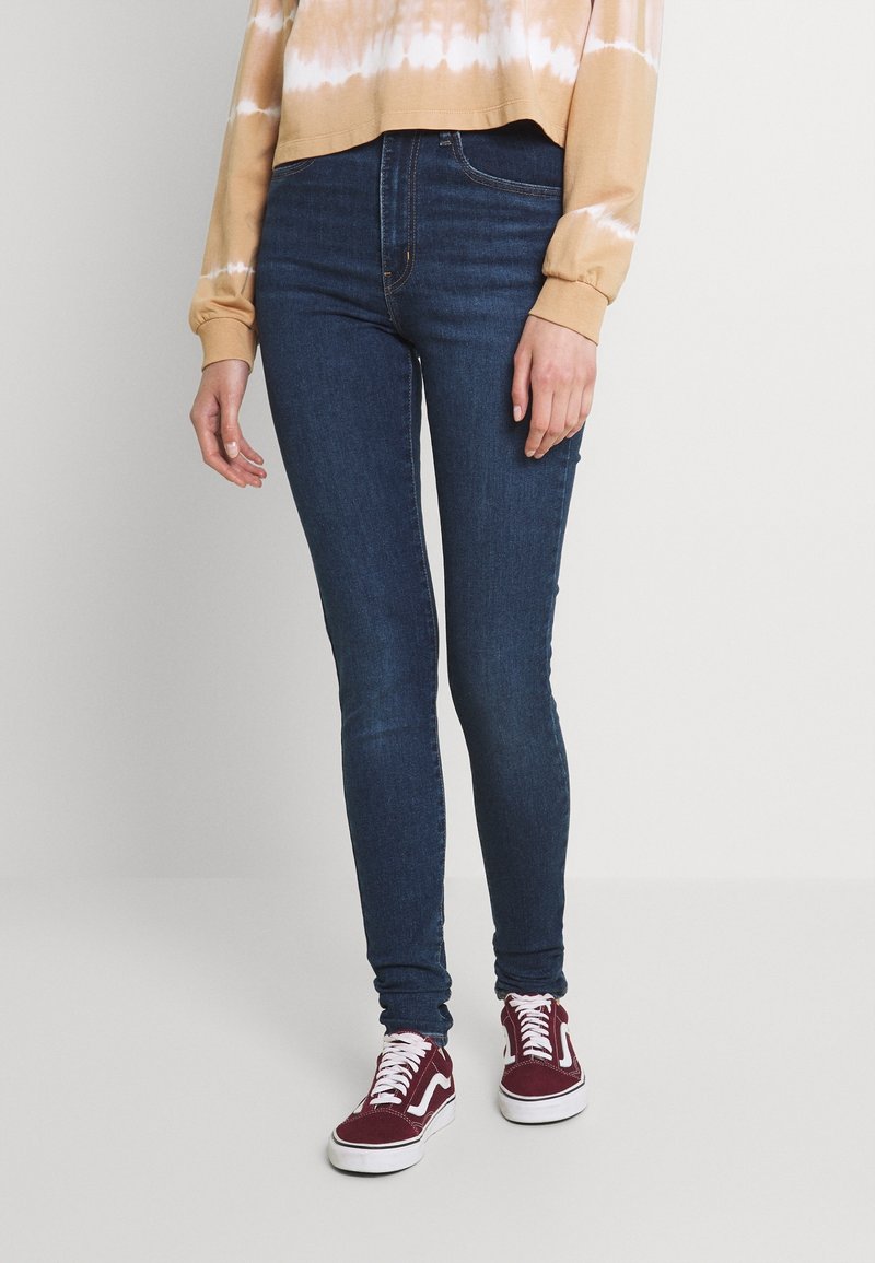 Jean skinny en denim bleu foncé, avec un design classique à cinq poches. Assorti à des baskets bordeaux et un sweat-shirt tie-dye beige.
