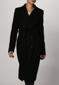 Sort uld trenchcoat med dobbeltknap, bælte i taljen, klippet krave og to forlommer. Glat tekstur, knælang design.