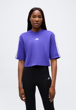 Mujer con una camiseta corta morada de Adidas y mallas negras de Adidas, de pie frente a un fondo blanco liso.