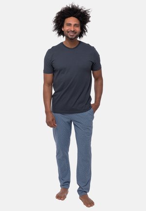 Mann mit lockigem Haar lächelt, trägt ein dunkelgraues T-Shirt und gemusterte blaue Pyjamahosen, steht barfuß vor einem weißen Hintergrund.