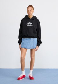 Čierna mikina s kapucňou a bielym logom, skombinovaná s džínsovou sukňou. Červené tenisky s bielymi šnúrkami a svetlo šedé ponožky dopĺňajú outfit.