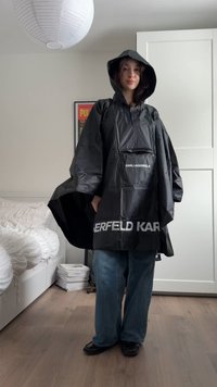 Persoon draagt een zwarte Karl Lagerfeld regenponcho met capuchon, blauwe spijkerbroek en zwarte schoenen, staand in een slaapkamer met witte meubels.