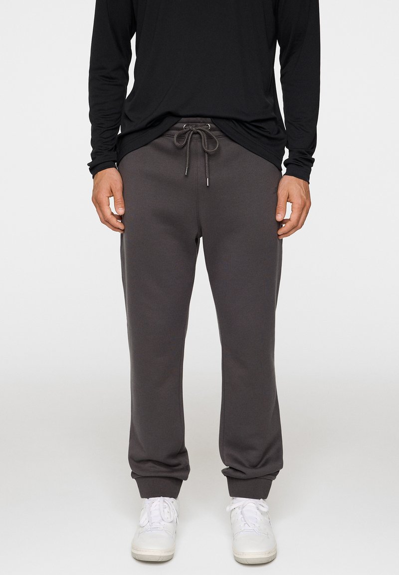 Szare spodnie dresowe z elastycznym pasem z troczkiem, elastycznymi mankietami i gładką fakturą, w zestawie z czarną koszulą z długim rękawem oraz białymi sneakersami.