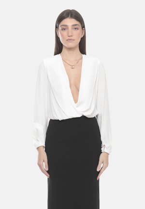 Donna con capelli lunghi e scuri che indossa una camicetta bianca con scollo a V profondo e una gonna nera a vita alta, in piedi davanti a uno sfondo bianco uniforme.