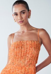 Oranje jurk met geplooide textuur en dunne bandjes. Heeft een bloemenpatroon en een getailleerde corsage. Gouden creolen maken de look compleet.