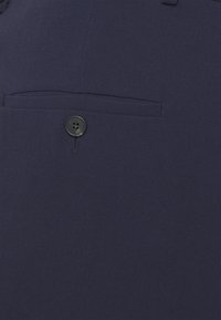 Tessuto blu navy con una tasca frontale e dettaglio del bottone nero. La texture appare liscia, adatta per abbigliamento formale.