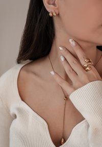 Goldene Tropfenohrringe, geschichtete goldene Anhänger-Halskette und ein spiralförmiger Goldring. Das Model trägt einen weißen gerippten Pullover und hat manikürte Nägel.