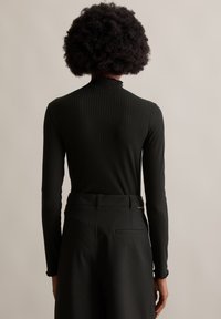 Schwarzes, geripptes Langarmoberteil mit einem Rüschensaum, kombiniert mit hoch taillierten schwarzen Hosen mit feinem Streifenmuster und einer Gesäßtasche.