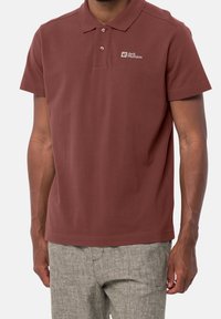 Polo bordeaux à col avec patte à trois boutons et logo brodé. Texture douce et lisse, associé à un pantalon gris clair.