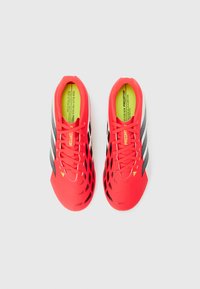 Κόκκινα και μαύρα ποδοσφαιρικά παπούτσια με κορδόνια, μαύρες ρίγες στα πλαϊνά και κίτρινες εσωτερικές σόλες με κείμενο, σε θέα από πάνω σε λευκό φόντο.