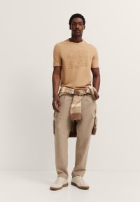 Beige t-shirt with embossed text, light brown high-waisted trousers, and off-white sneakers. A multi-colored plaid sweater tied around the waist.