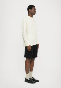 Kremfarget lin skjorte med lange ermer og mandarin krage, i kombinasjon med svarte shortser og svarte loafers med beige sokker.