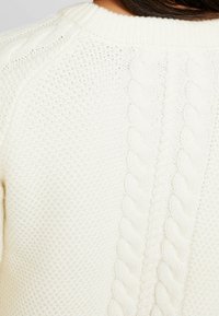 Frau in einem weißen, gestrickten Pullover mit Kabel- und Strukturmustern, der den Halsausschnitt und den Oberkörper zeigt.