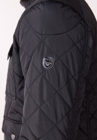 Manche de veste matelassée noire avec couture en forme de losange et une petite étiquette de marque "Pier-One" près de l'épaule.