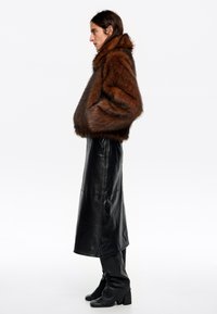 Brązowa kurtka z sztucznego futra z wysokim kołnierzem, noszona na długiej czarnej skórzanej spódnicy i czarnych kozaczkach do kolan. Modelka stoi w profilu.