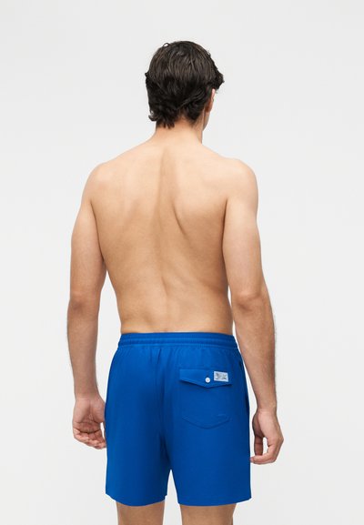 Mężczyzna stojący tyłem, ubrany w jasnoniebieskie kąpielówki z tylną kieszenią i zapięciem na guzik, na jednolitym jasnym tle.