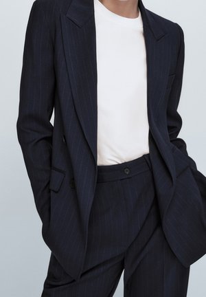 Personne portant un costume à rayures foncées avec une chemise blanche, mains dans les poches, sur un fond gris clair.