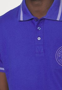 Fialové polo tričko s texturovanou tkaninou, vybavené dvoubarevnými knoflíky, kontrastními světle modrými pruhy na límci a detailním kulatým logem.